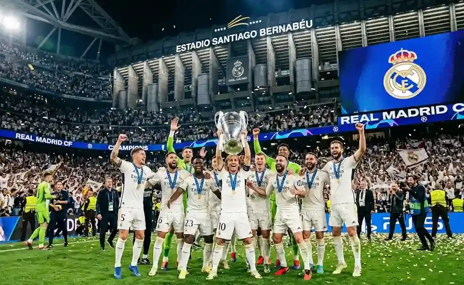 Real Madrid players celebrating with the UEFA Champions League trophy on the pitch at Santiago Bernabéu stadium under glowing stadium lights.