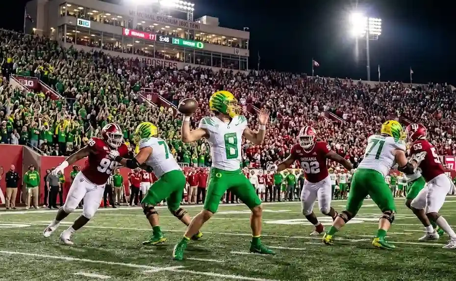 Oregon Ducks quarterback in a green and yellow uniform winding up to throw a pass during an intense Big Ten college football game against the Indiana Hoosiers under bright stadium lights.
