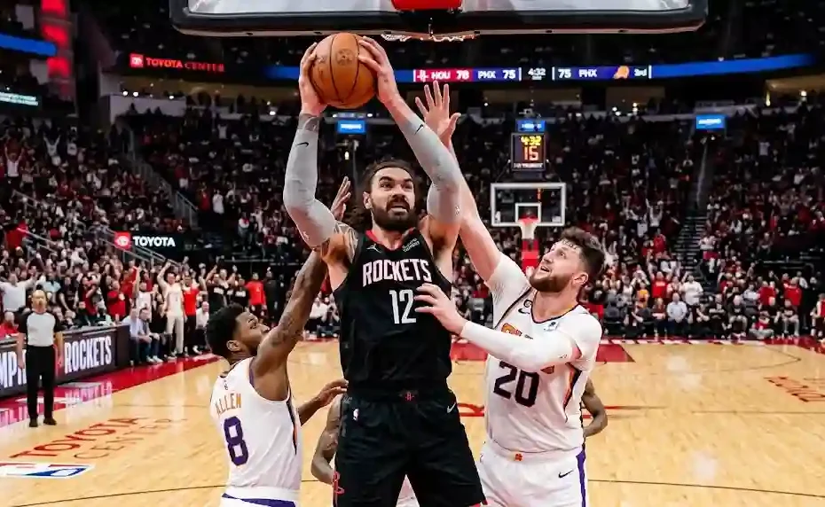 NBA center Steven Adams in a black Houston Rockets jersey grabbing a strong two-handed offensive rebound in the paint surrounded by defenders under bright arena lights.